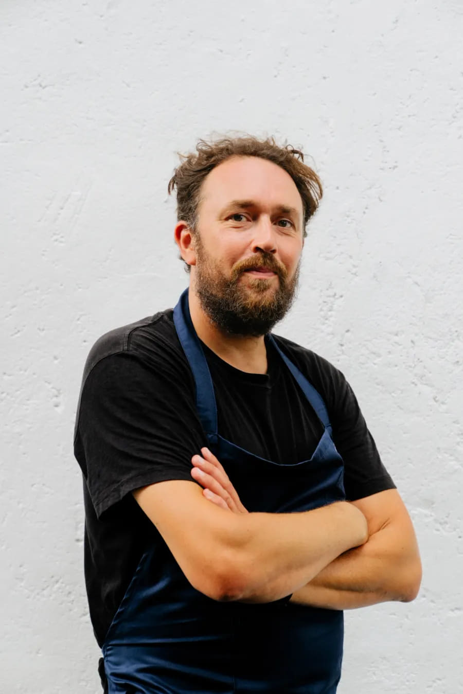Cameron Emirali in a dark shirt and navy apron, arms crossed, against a white wall