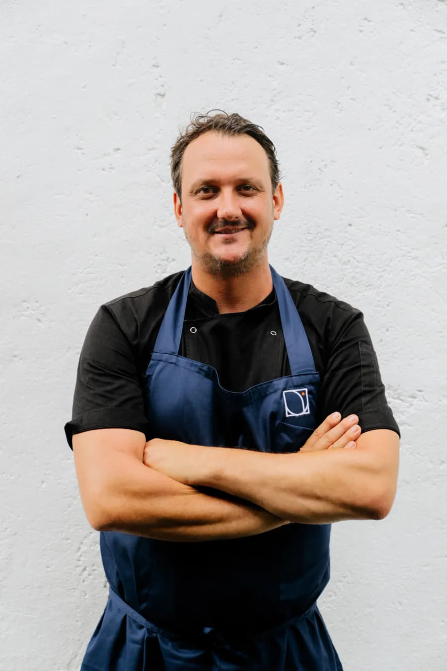 Kieran Morland in a dark shirt and navy apron, arms crossed, against a white wall