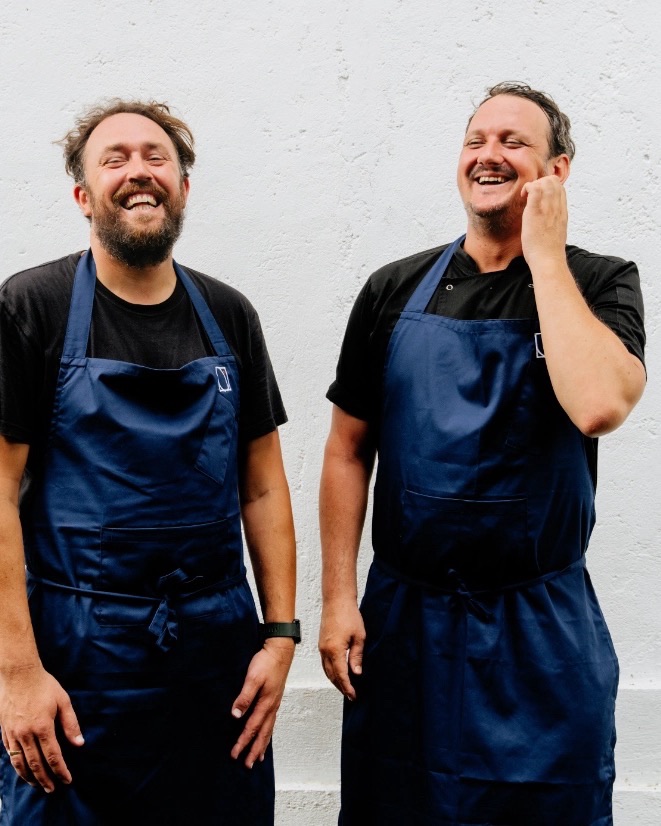 Cameron Emirali and Kieran Morland laughing together in navy aprons against a white wall