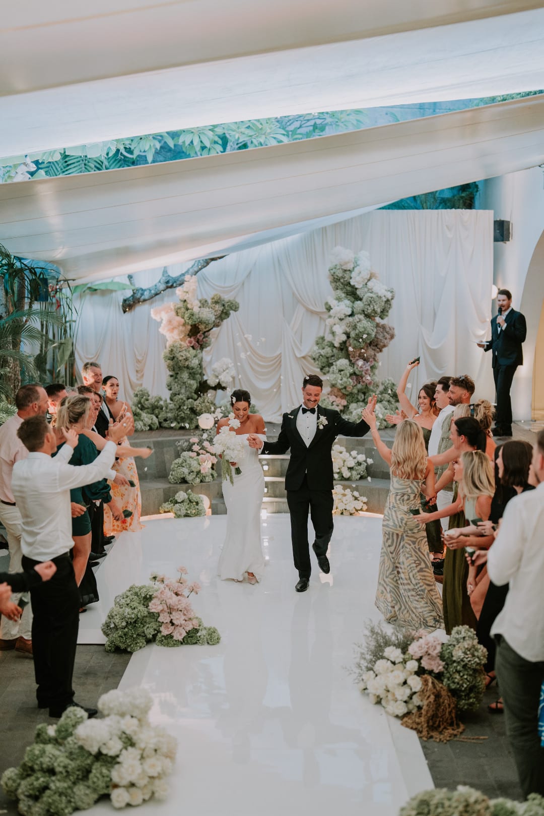 A groom walking between standing guests under a white floral arch at Luma