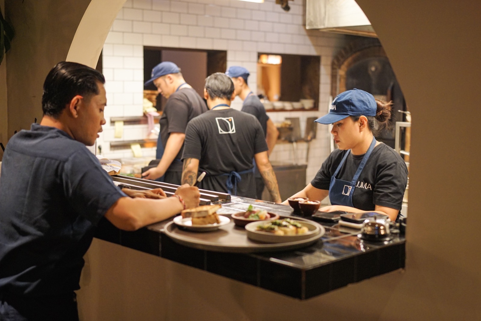A waiter collecting plates from the pass mid-service while the kitchen works behind the arch